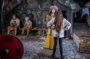 A young visitor in modern clothing stands with hands on hips, facing a Viking woman in a fur hat inside a reconstructed Viking village.
