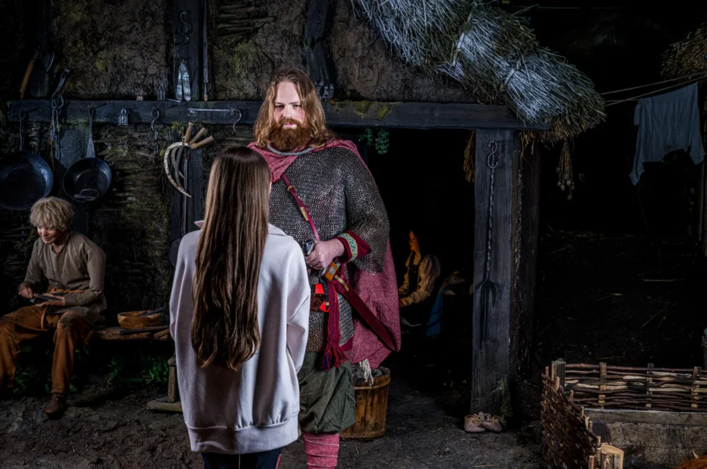 A young visitor in modern clothing stands, facing a warrior in chainmail with his hand on a sword inside a reconstructed Viking village.