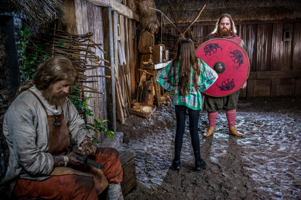 A young visitor in modern clothing stands with hands on hips, facing a Viking warrior in chainmail with a red shield inside a reconstructed Viking village.