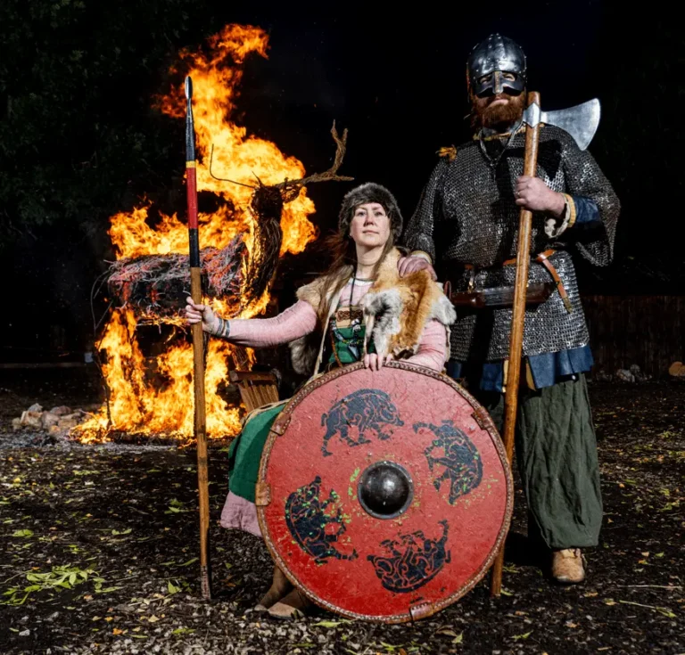Viking reenactors in traditional attire standing in front of a burning stag effigy, with a woman holding a shield and spear and a man in chainmail holding an axe.
