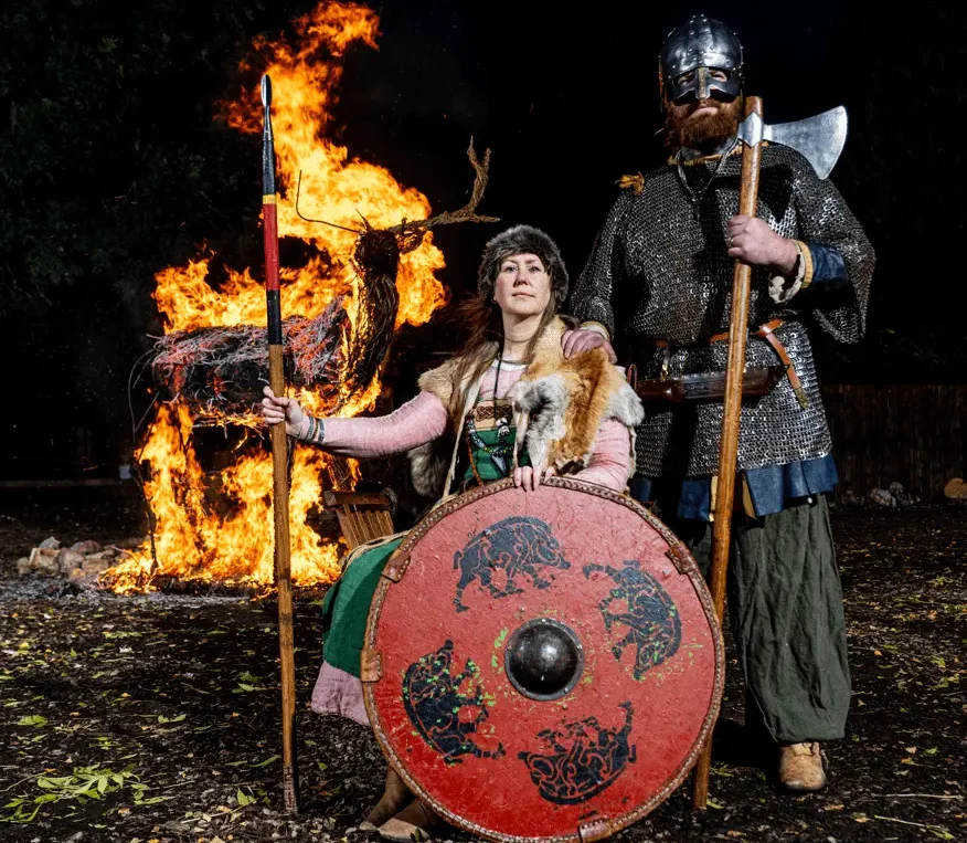 Viking reenactors in traditional attire standing in front of a burning stag effigy, with a woman holding a shield and spear and a man in chainmail holding an axe.
