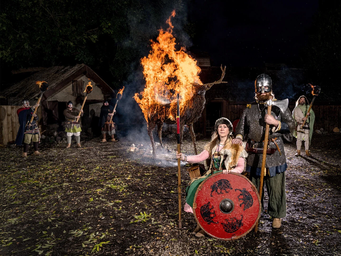 A group of Viking reenactors standing near a burning wooden stag effigy during a night event, showcasing traditional Viking attire and weapons.