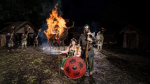 A group of Viking reenactors standing near a burning wooden stag effigy during a night event, showcasing traditional Viking attire and weapons.