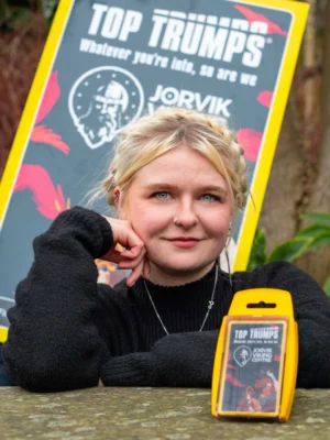 Katie Smith, an illustrator with blonde hair in a braided style, leans on her hand in front of a stone surface, next to a deck of JORVIK-themed Top Trumps cards in yellow packaging. Behind her, a sign with 'Top Trumps' branding and a Viking illustration is visible.